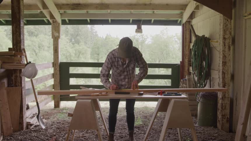 A woman in a plaid shirt and cap measures a long wooden board on sawhorses inside a rustic opensided workshop