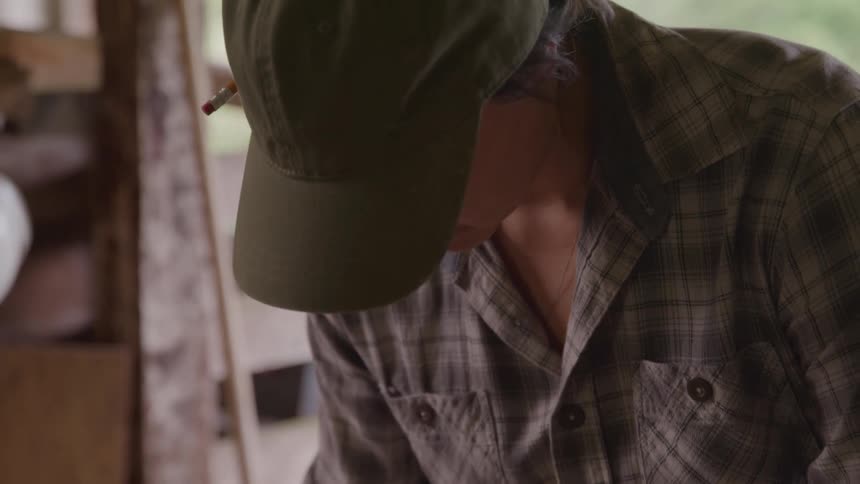 A woman wearing a hat works with wood inside a shed at midday