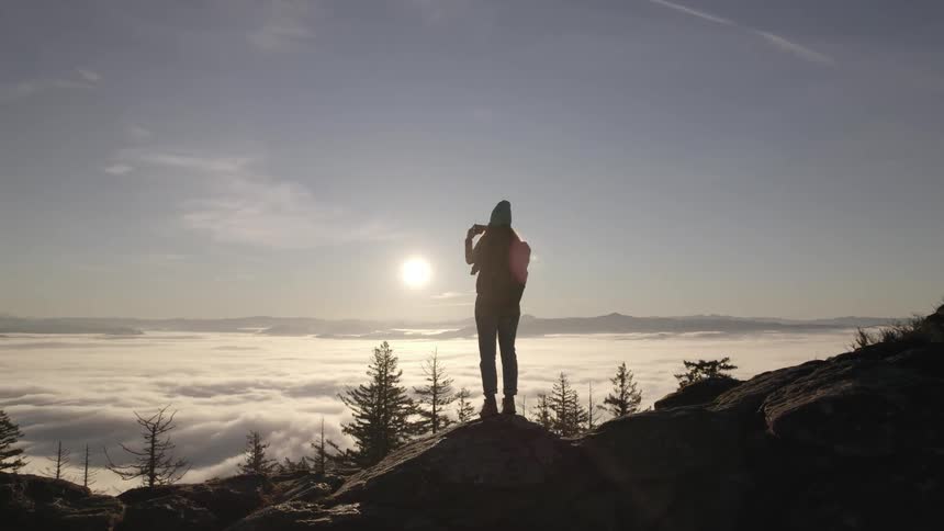 A young woman hiking in the woods stops on a rocky hill to take pictures of nature with a series of trees in the distance and the setting sun in the sky as the backdrop