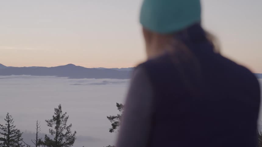 Person with a beanie looks at the mountainous horizon at sunrise above the clouds