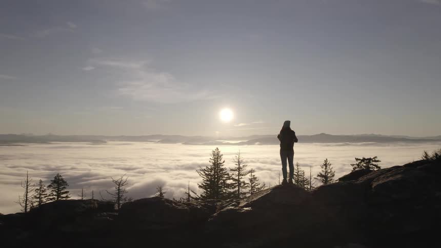 A person is silhouetted against a bright hazy sky standing on a rocky mountain ridge below them a thick blanket of clouds stretches across the landscape with other distant mountains visible in the background the sun shines brightly through the haze