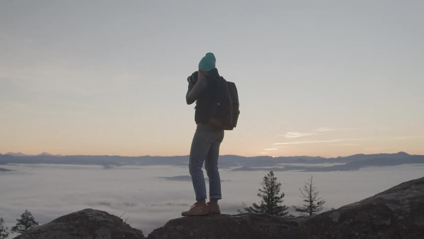 A young woman wearing a sweatshirt backpack jeans and sneakers stands on a mountaintop taking photographs of nature at sunset