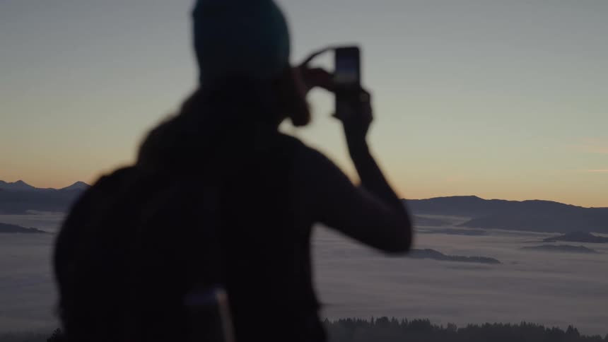 A person is standing on a mountain peak taking a photo of a sunrise or sunset with their phone the view shows a landscape of mountains covered in a blanket of fog or clouds