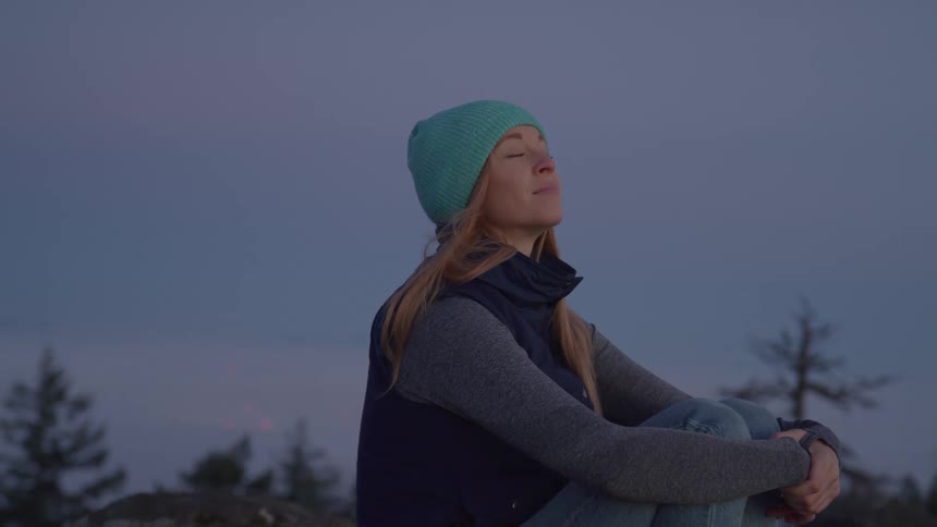 A contemplative woman sits peacefully in a natural setting at twilight looking up at the sky