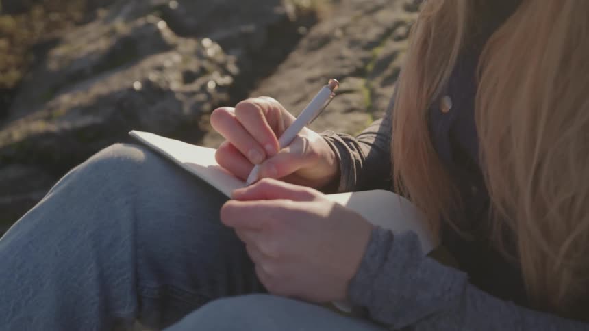 A young woman sits on a rock writing with a pen in a notepad at midday
