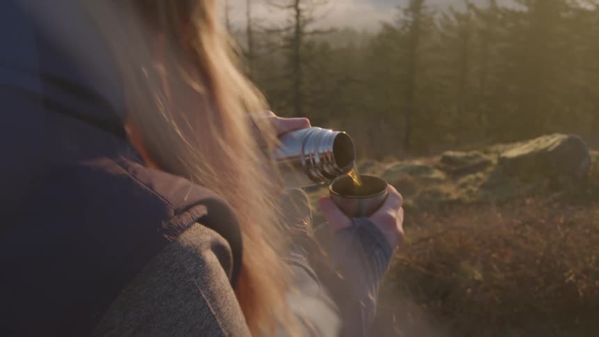 Person pouring a drink from a thermos into a cup outdoors during sunset