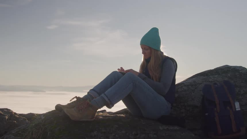 A young woman sits on top of a rock writing in her journal while wearing a hat jeans and boots with the setting sun in the sky