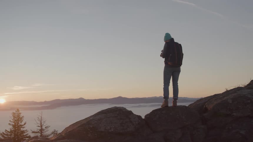 A young woman walks from one rock to another on a mountain preparing to take a picture with her digital camera of the setting sun against the distant mountain range