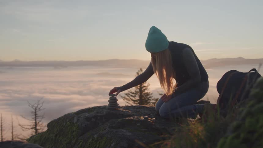 Female backpacker kneels on the stony ground overlooking a foggy lake while stacking stones