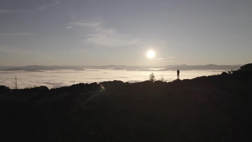 A person hiking stands on the peak of a mountain with the orange setting sun illuminating the surroundings