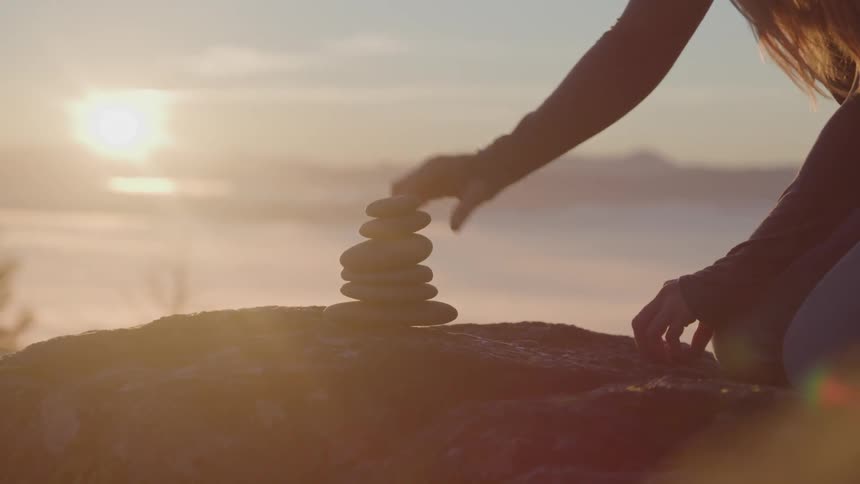 A young girl on a mountain stacks rocks into a pyramid on a larger rock with the orange sky and setting sun in the background
