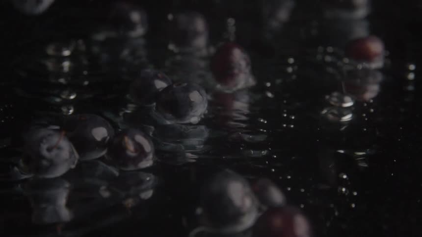 A shot shows water pouring down splashing into a pile of darkcolored grapes the grapes and the water are on a black surface creating a highcontrast and dramatic effect