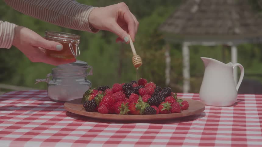 A persons hands drizzle honey over a platter of fresh strawberries and blackberries on a checkered picnic tablecloth outdoors