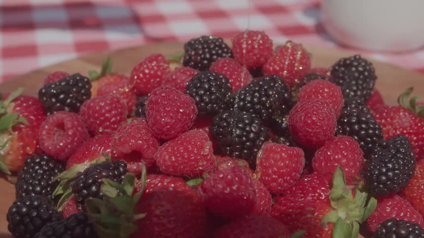A variety of berries including raspberries and blackberries being drizzled with syrup under the sun