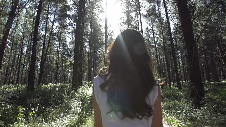 A young woman with long brown hair and dressed in a white dress walks through the forest