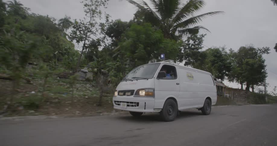 White van driving down a street in a tropical area with lights blinking