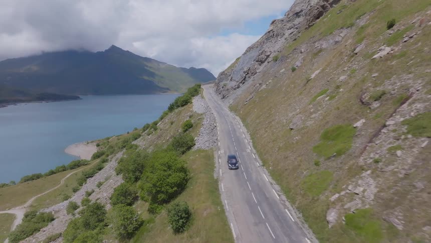 A car drives along a mountain road with a large tranquil blue lake on one side and a rocky hillside on the other the sky is partly cloudy and the overall scenery is lush and green