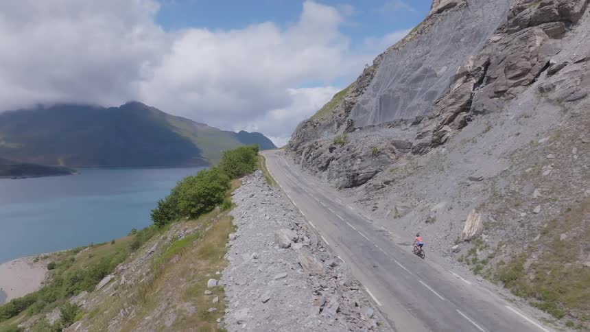 Cyclist rides along a narrow road beside a lake and rocky slope