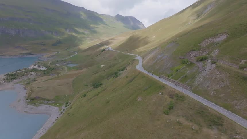 Cyclists riding on a winding mountain road alongside a large lake surrounded by vast green hills