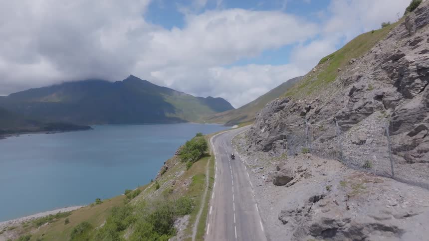 A highangle drone shot shows a motorcyclist riding along a winding road beside a large lake in a mountainous landscape the road curves around the base of a steep rocky mountain while the bluegreen water of the lake stretches out to the left
