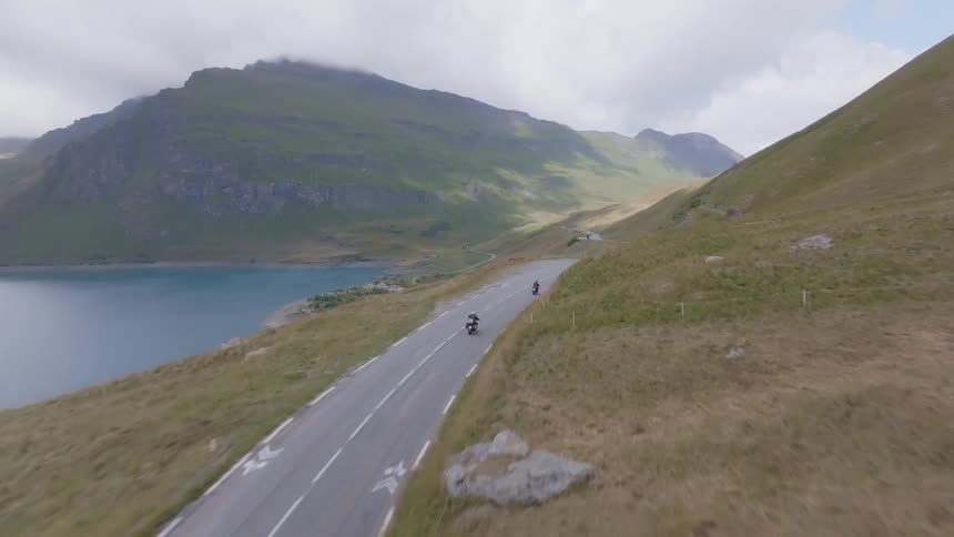 A motorcyclist rides along a winding road next to a large blue lake with a steep green mountain slope rising on the other side the camera follows the motorcycle as it travels along the road in a scenic outdoor setting