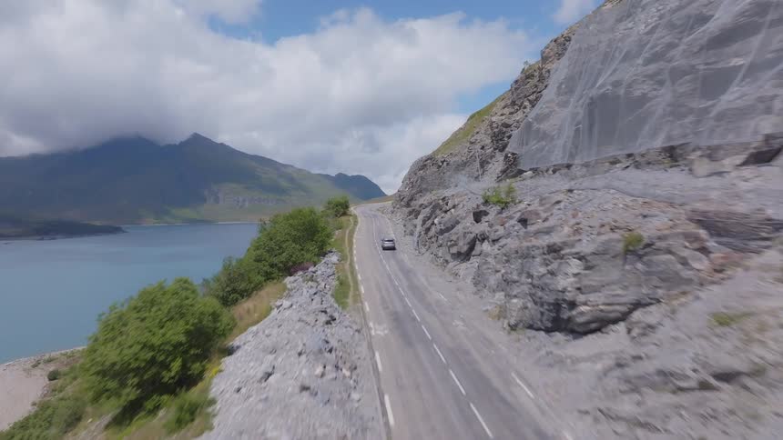 A car drives along a road built on the side of a mountain by the coast at midday