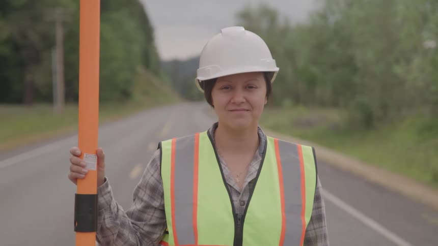 A young woman holds a traffic sign rerouting traffic due to construction wearing a hard hat and green vest on the freeway at midday