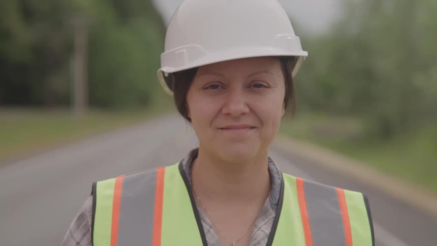 A young woman wearing a hard hat and green vest stands in the middle of the road at midday