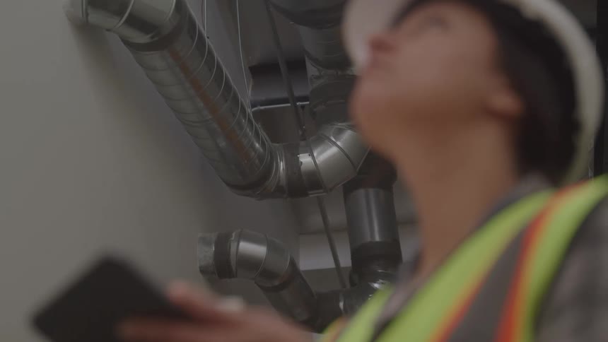 Woman wearing a helmet and reflective vest holding a tablet while inspecting ceiling piping
