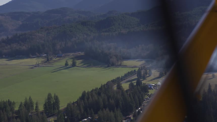 An aircraft flies over a rural community surrounded by green vegetation at midday