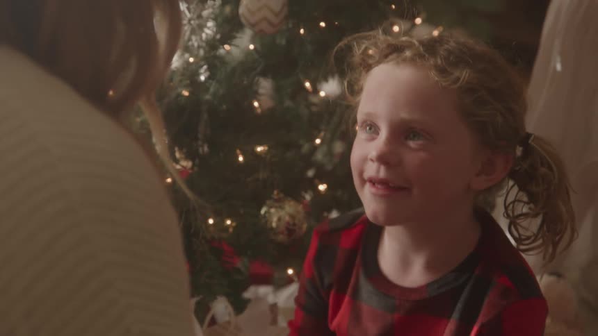 The video shows a young girl with curly red hair wearing red and black plaid pajamas looking up and smiling at someone offcamera she is sitting in front of a brightly lit and decorated christmas tree the scene has a warm festive and cozy atmosphere
