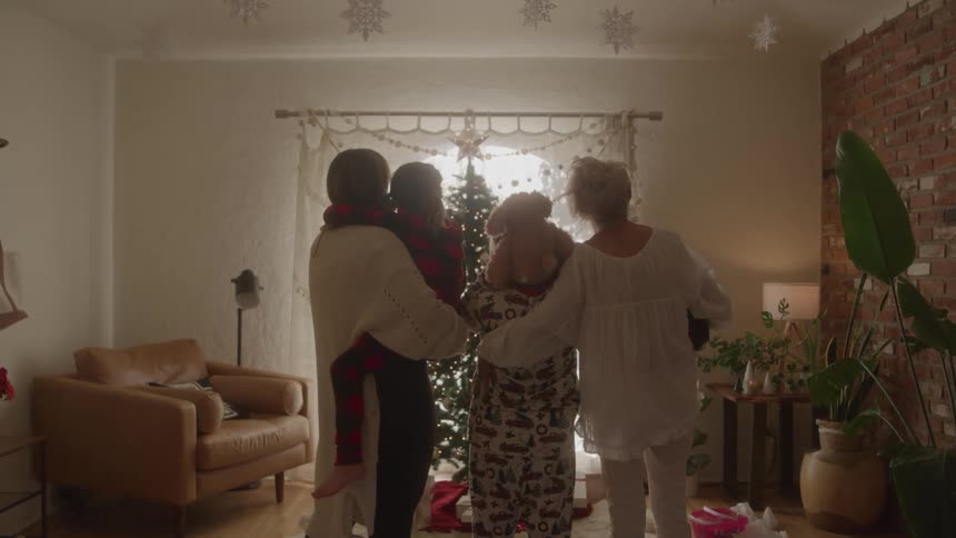 A group of three adults are standing with their backs turned in a living room and one of them is holding a child on her lap