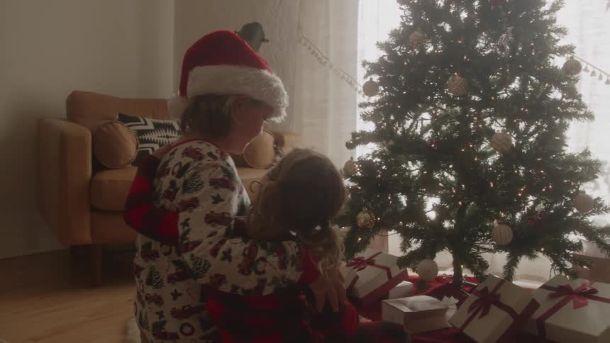 A mother and child sit beside a christmas tree wearing pajamas and looking excitedly at the christmas presents at midday