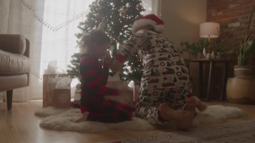 A parent and a child both wearing christmas pajamas and santa hats are seen walking toward a christmas tree they kneel in front of the tree which is decorated with lights to open presents