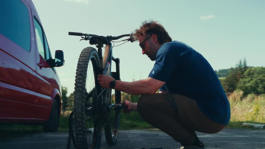 A man wearing sunglasses kneels beside a mountain bike making adjustments to its front wheel near a red van
