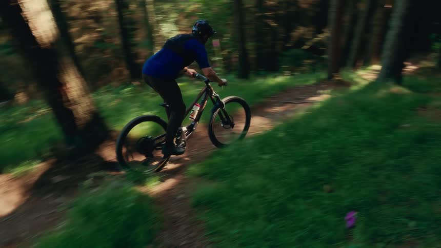 The video shows a person riding a mountain bike downhill on a narrow winding trail through a forest the camera follows the biker from the side as they ride past trees and a low stone wall