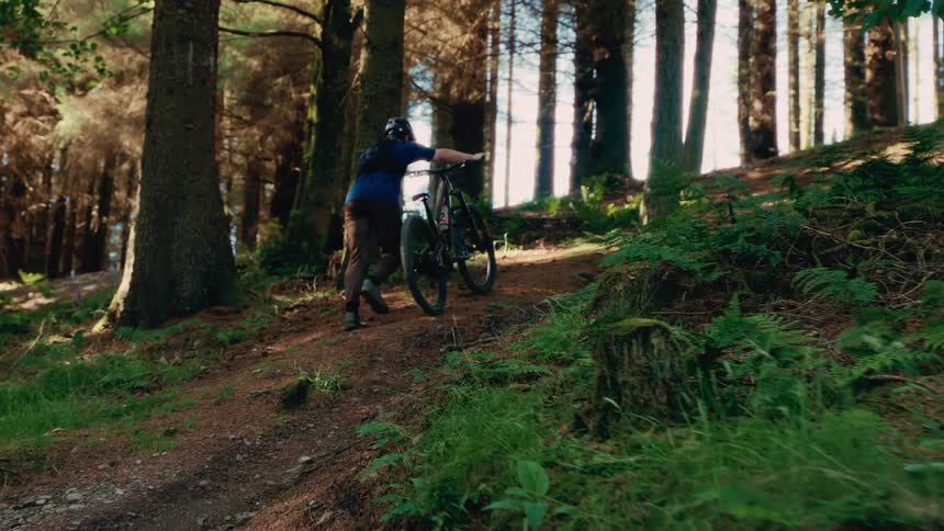 The video shows a person pushing a mountain bike uphill on a dirt trail through a forest the sun is shining through the trees casting long shadows