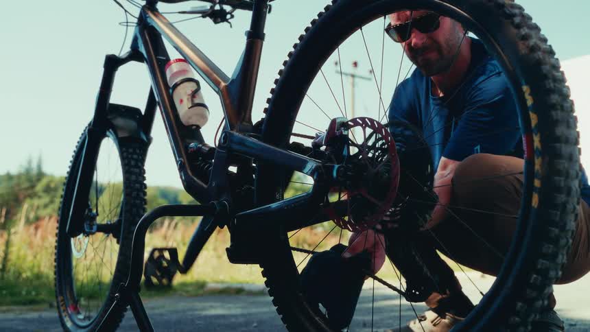 A man wearing sunglasses crouches beside a mountain bike adjusting the rear wheel mechanism on a sunny day