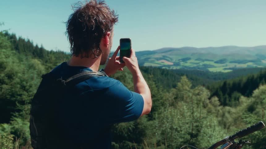A man sits on a mountain bike taking a photo of the expansive green valley and rolling hills before him