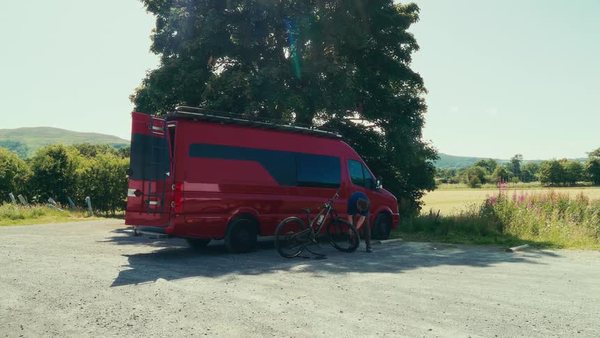 The video shows a person using a bike pump to inflate a tire the person is standing next to a large red camper van that is parked under a tree a field is visible in the background and the video has a filmsupply watermark