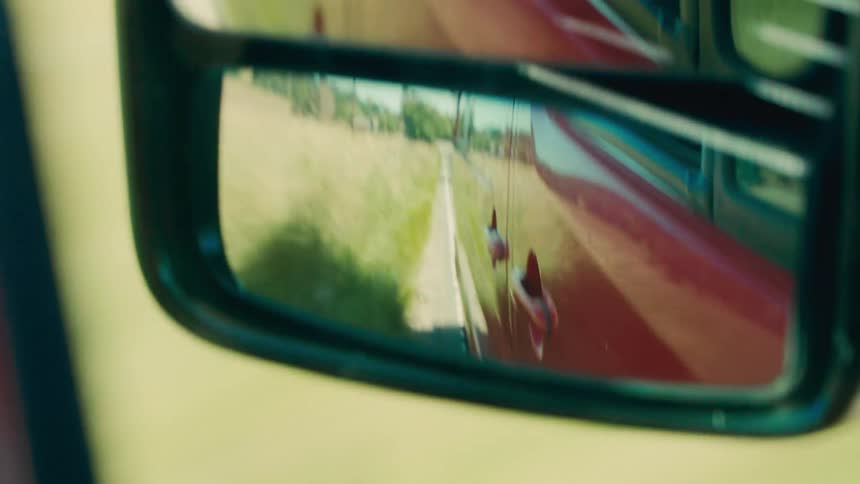 POV looking into the driver's side mirror of a red van driving on a rural road as the lush countryside goes by in the mirror's reflection.