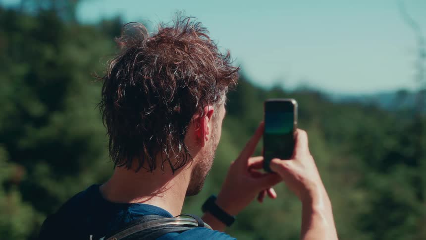 The video shows a man from behind holding a smartphone up to take a picture of a scenic view the man has brown hair and is wearing a blue shirt and a backpack the video has a watermark that reads filmsupply