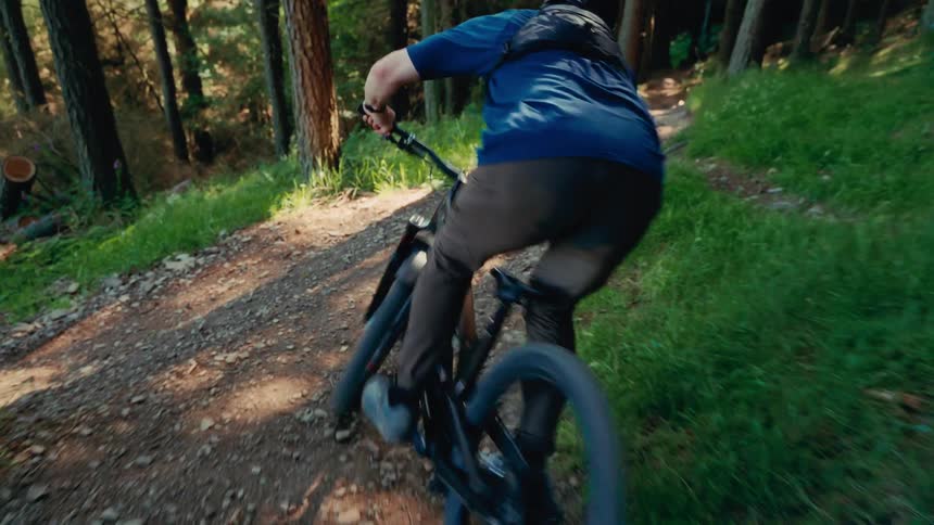 The video shows a person riding a mountain bike downhill on a dirt trail through a dense forest the camera is stationary and captures the person as they ride past a tree and down the path
