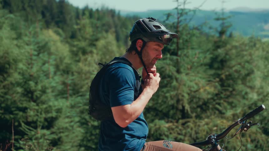 A mountain biker with a blue shirt and backpack stands in a forest putting on a helmet while holding onto the handlebars of their bicycle
