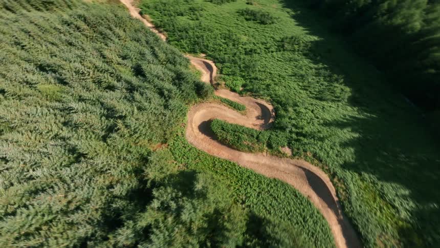 The video shows a winding dirt track presumably a mountain bike trail running through a densely forested area the camera moves in an aerial shot revealing a beautiful green hilly landscape with patches of trees and a bright blue sky in the distance