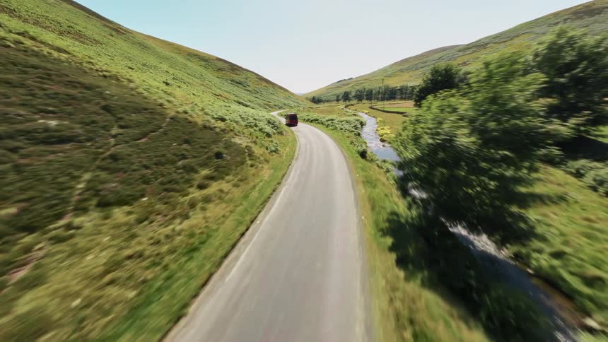 The video shows a red camper van driving down a road in a green hilly landscape the camera is positioned high above the road following the van as it drives the road has a small stream running parallel to it the video has a watermark that reads films
