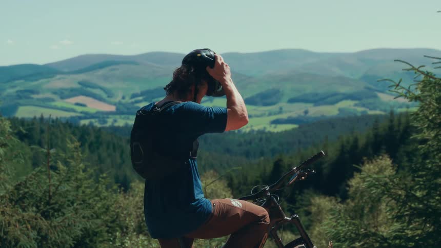 A mountain biker sits on his bike having removed his helmet looking down the forest path at midday