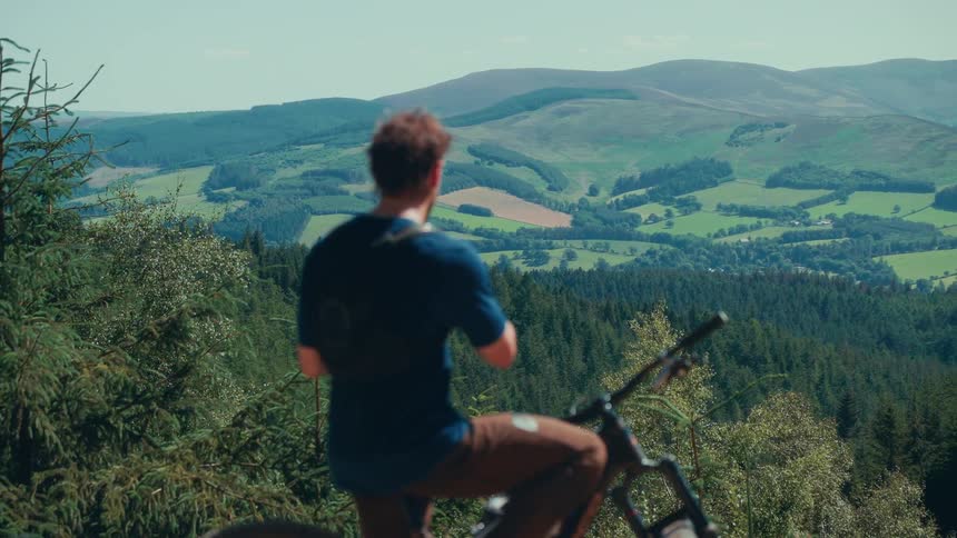 A man sits on a mountain bike taking pictures using his phone towards the expansive valley and rolling hills covered in forests and farmland