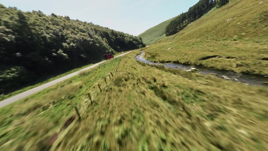A red van drives along a winding rural road beside a narrow stream in a lush green valley