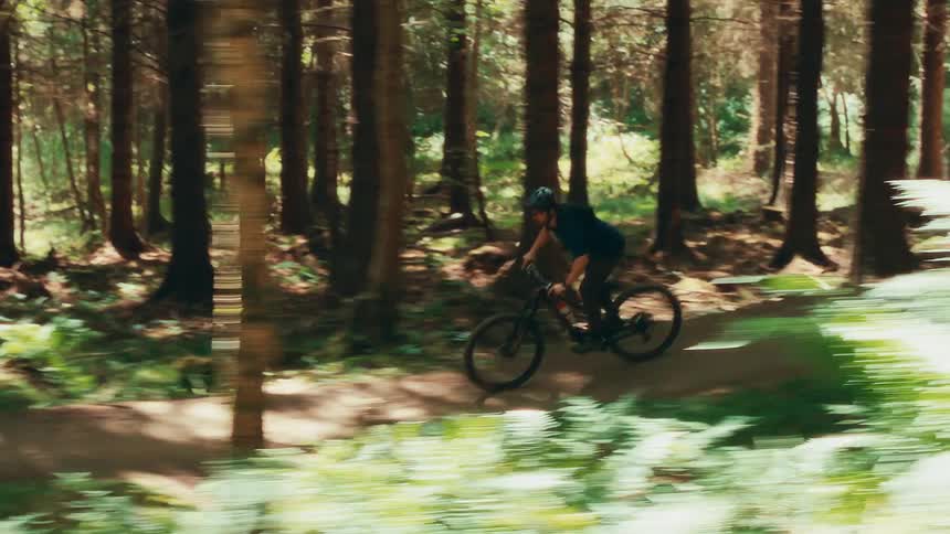 The video shows a person mountain biking through a sundappled forest the camera follows them as they ride along a winding dirt path weaving between tall trees the person is wearing a helmet and is focused on the trail ahead the watermark filmsupply